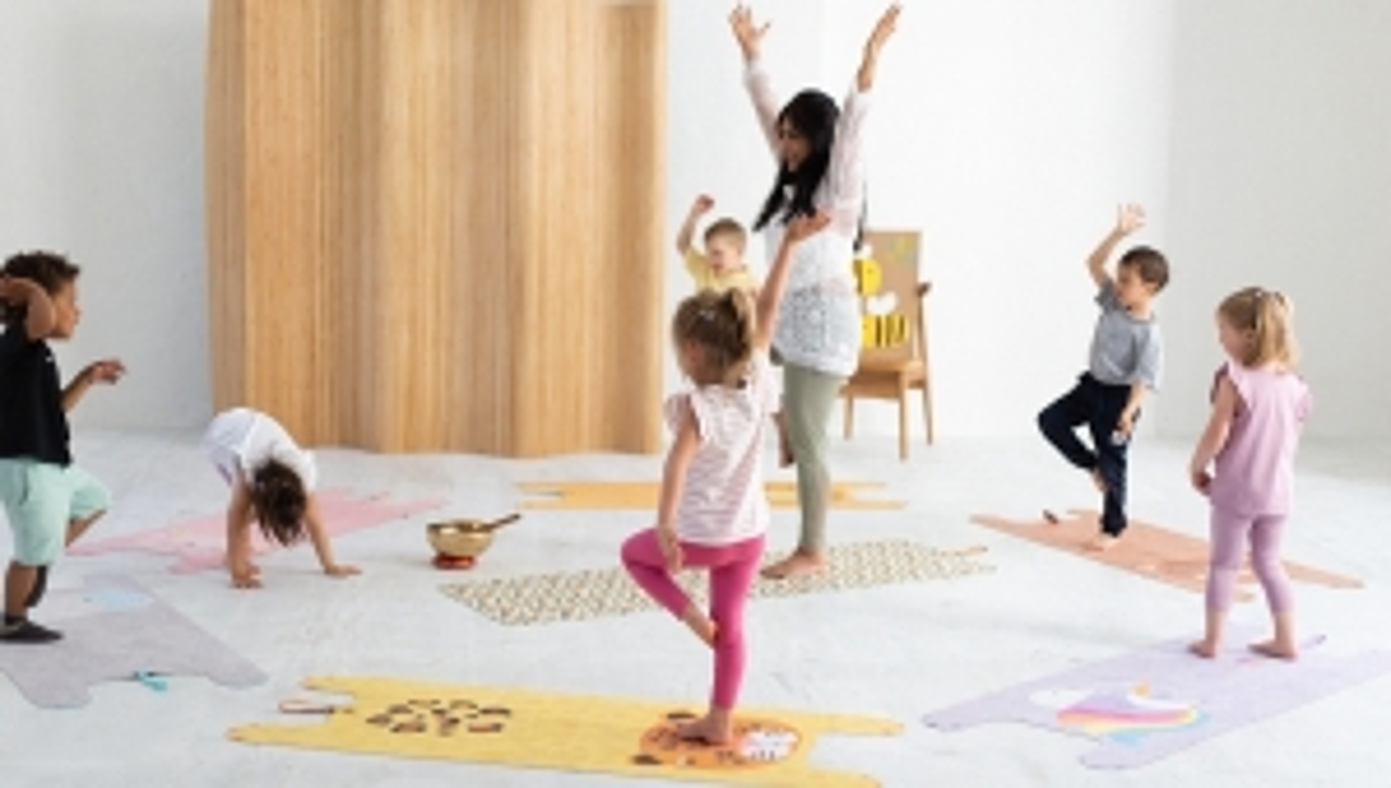© yogitier Kinder stehen im Kreis mit ihrer Betreuerin auf Yogamatten mit Tiermotiven und üben sich im Einbeinstand.